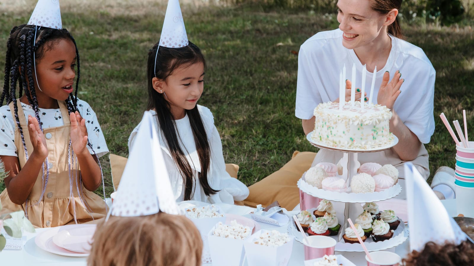 A delightful outdoor birthday party with kids and a woman celebrating with a cake and party hats.